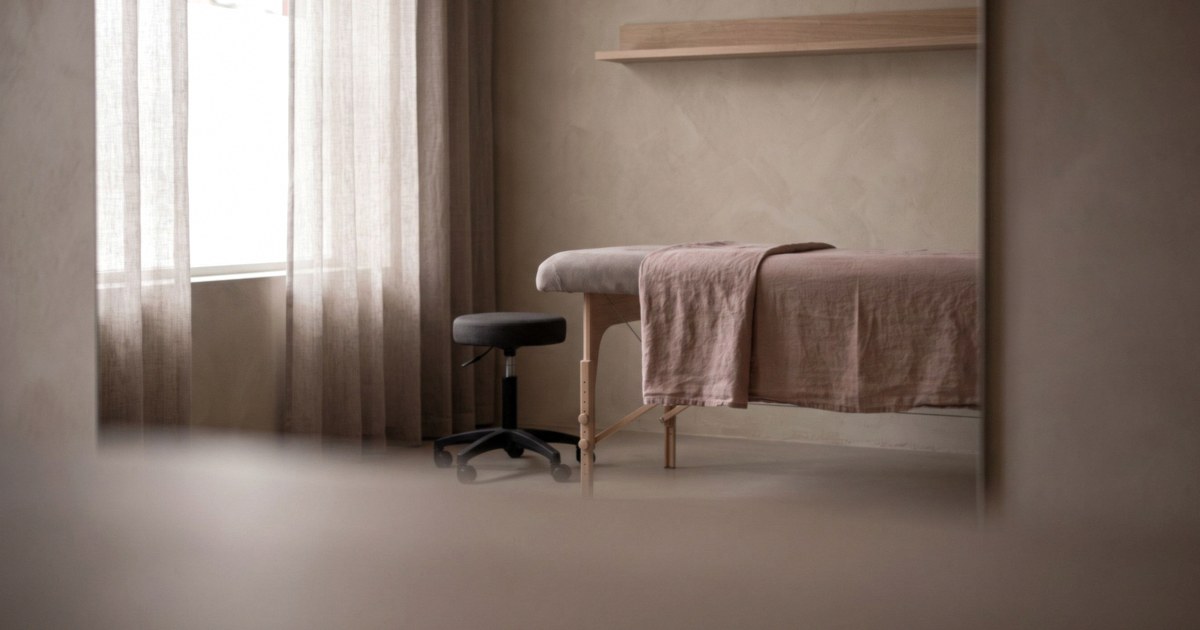 An empty treatment room with a linen-draped treatment bed and a rolling stool beside sheer curtains in soft daylight.