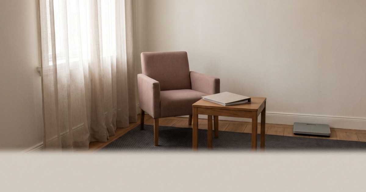 An empty consultation room with a dusty rose armchair, a wooden side table holding a closed binder and a floor scale by a curtained window.